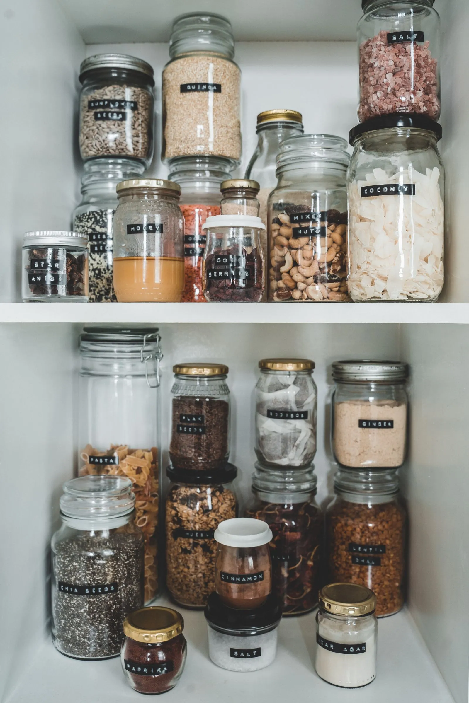 Labeled glass jars of pantry staples including cinnamon, salt, coconut, and mixed nuts arranged on white shelving