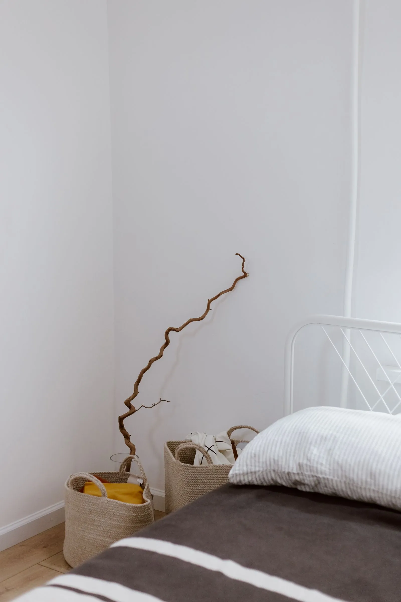 Two woven storage baskets on the floor in a bedroom corner, one holding a folded canvas tote bag and a folded yellow fabric item