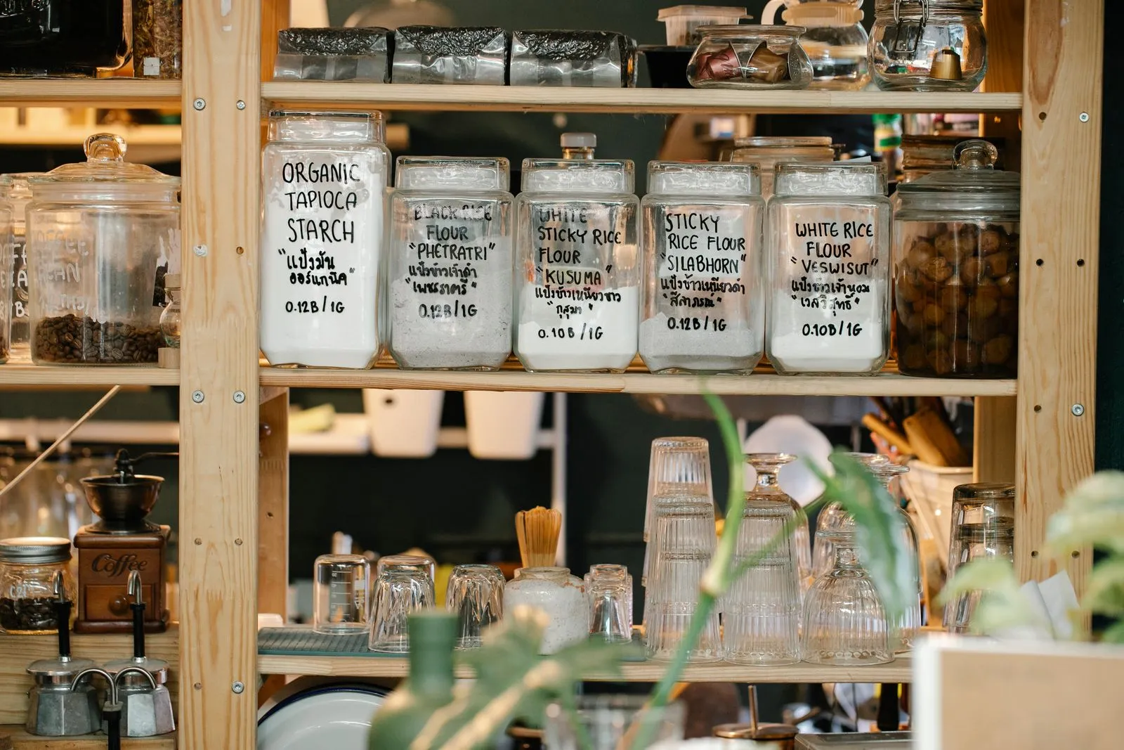 Individually labeled glass canisters of flour and starch lined up on a wooden pantry shelf, with jars of coffee beans and glassware nearby
