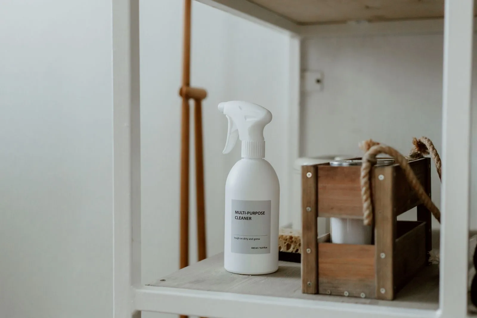 A spray bottle labeled multi-purpose cleaner on a cabinet shelf next to a small wooden crate, with a pipe running through the cabinet