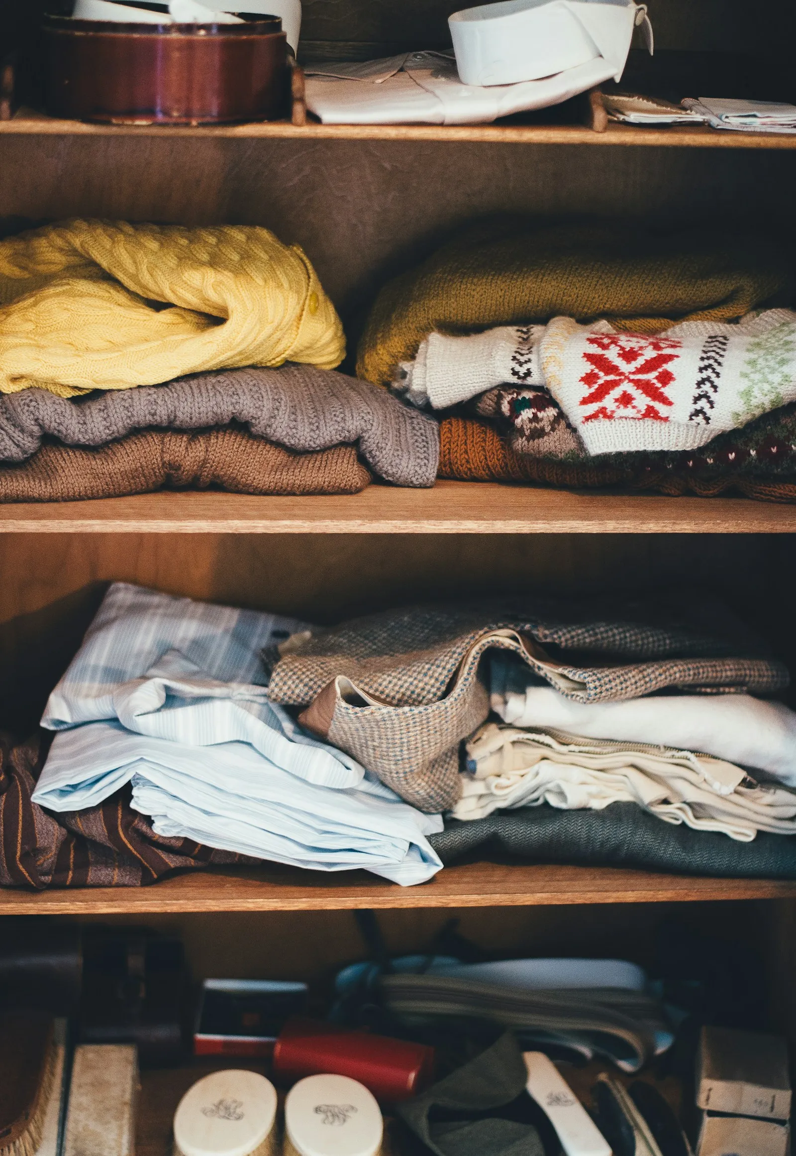 Wooden closet shelves stacked with folded knit sweaters and other folded clothing in a column of neat piles
