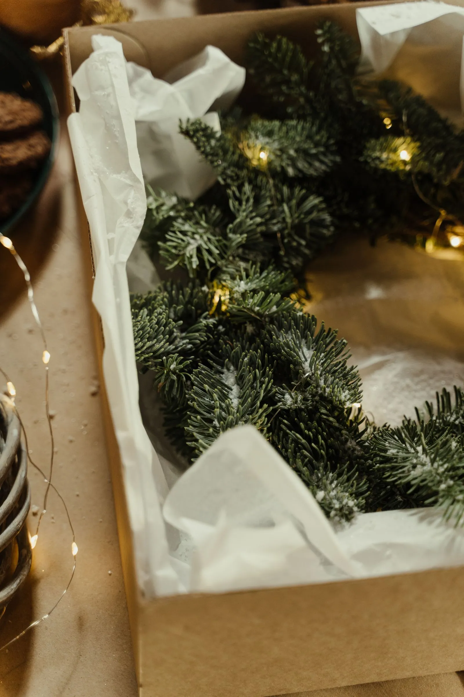 A wreath wrapped in white tissue paper inside a cardboard box, with a strand of warm string lights tucked among the branches