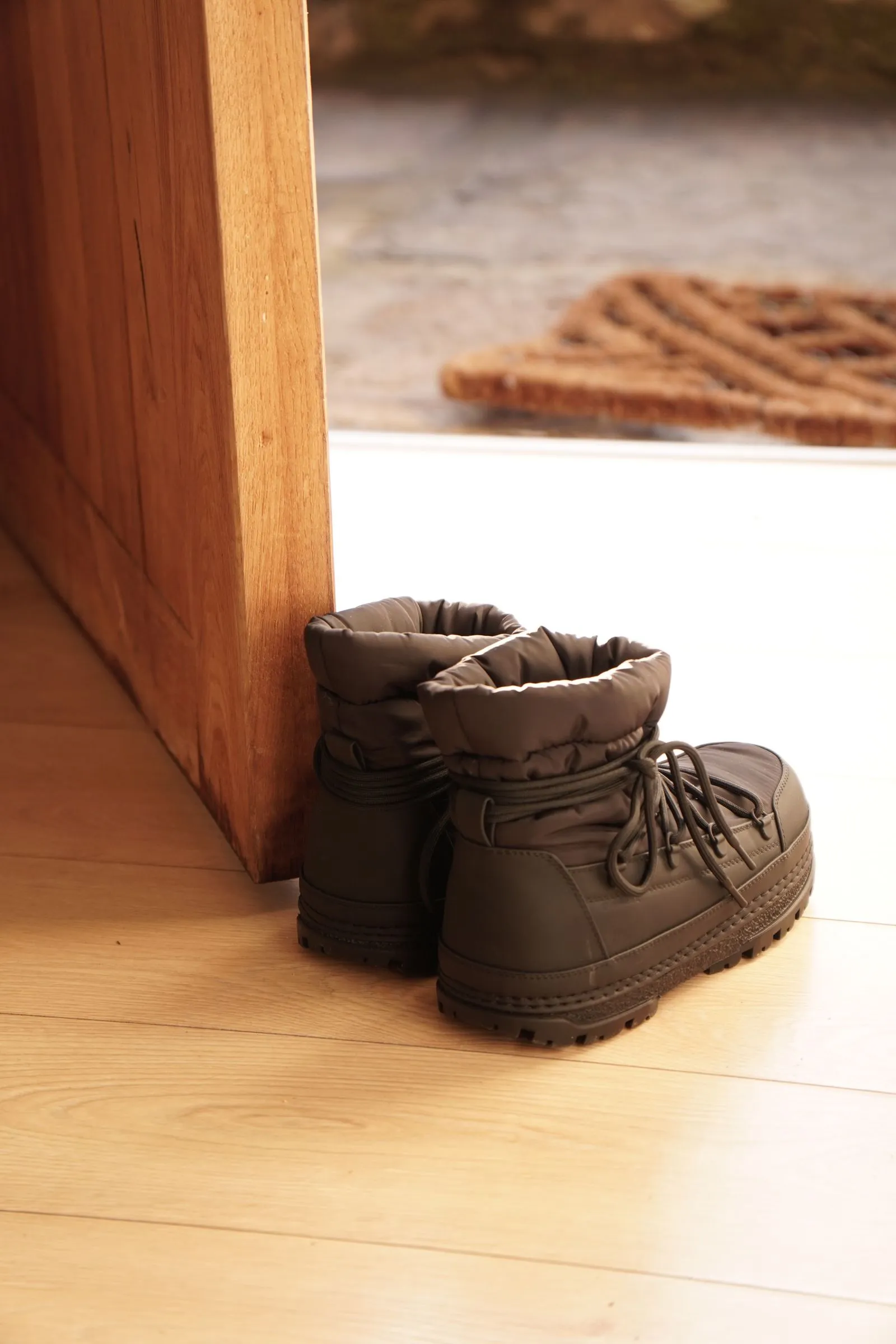 A pair of quilted olive-grey winter boots standing on a wood floor just inside an open front door, with a woven doormat visible on the step outside