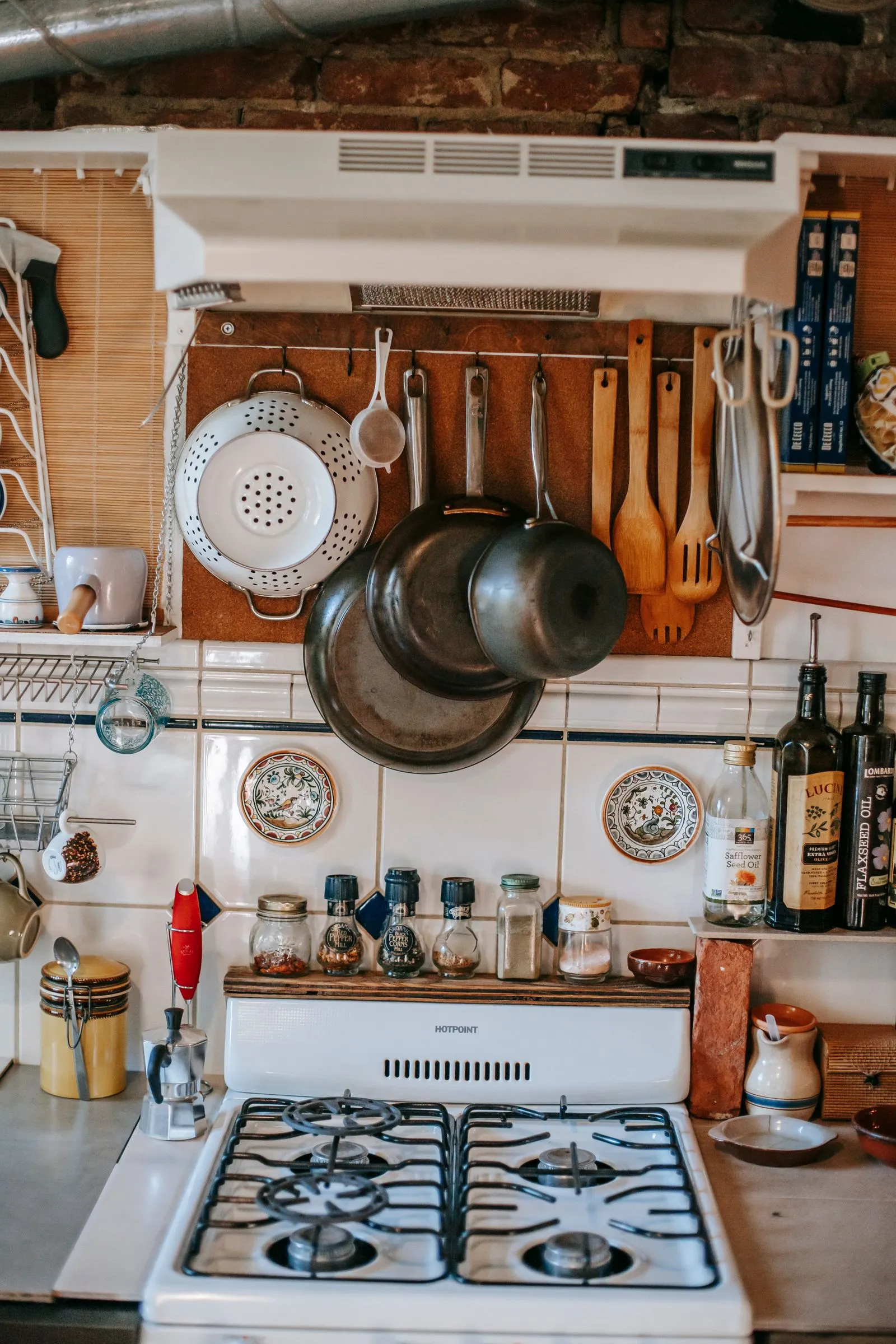 Wall-mounted hanging pots, a colander, and wooden utensils on hooks above a gas stove backsplash, with oil bottles on a shelf to the side